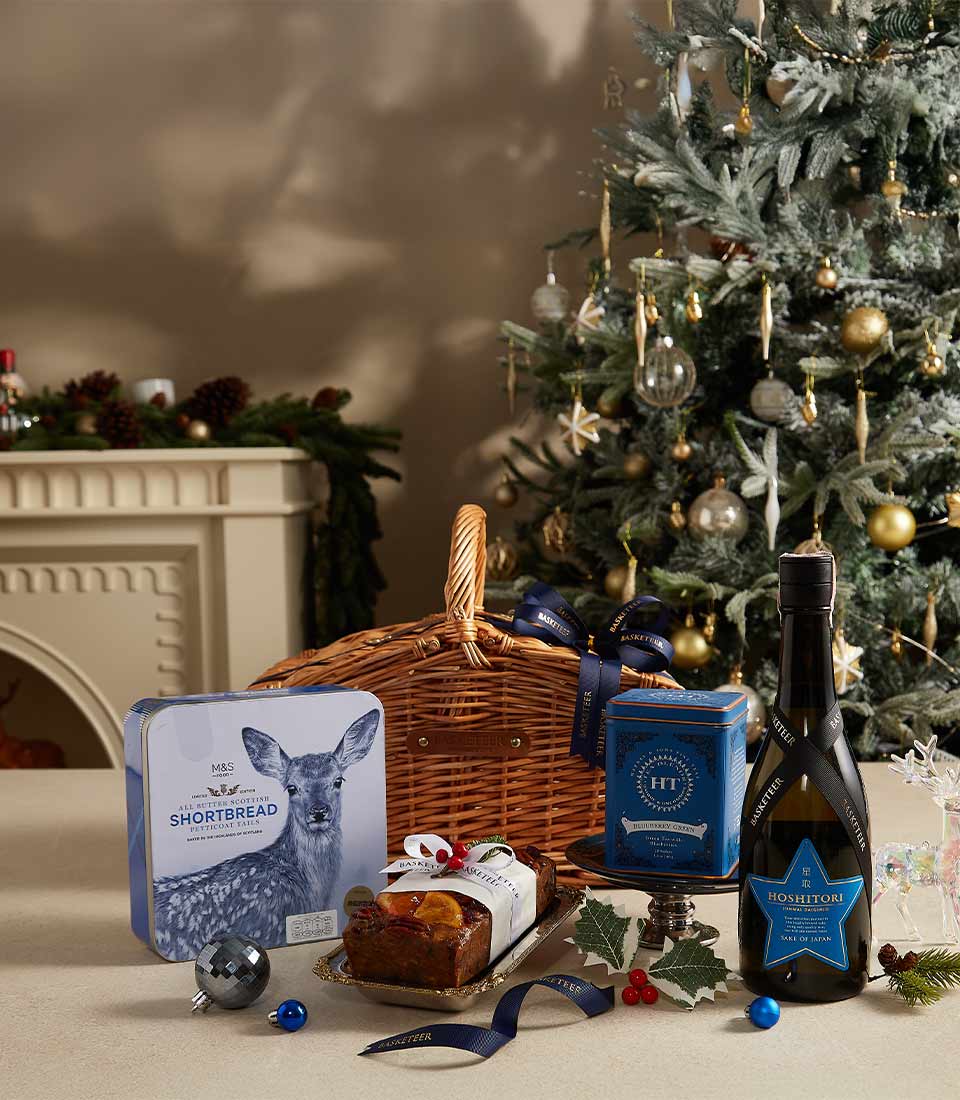 A wicker gift basket with navy ribbon filled with M&S shortbread, Harney & Sons tea, Hoshitori sake, Christmas fruitcake, and festive holiday decorations by a beautifully lit Christmas tree.