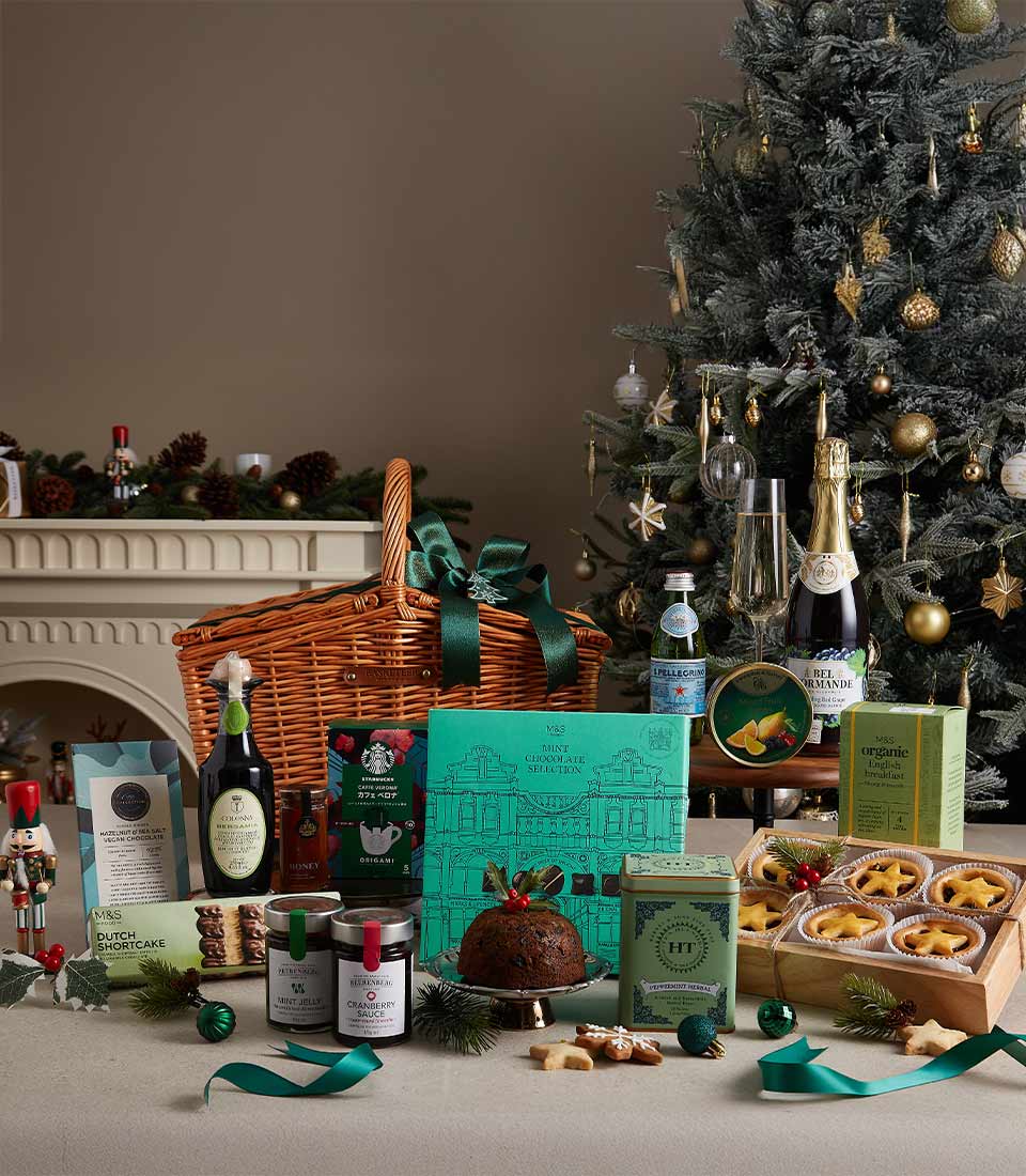 Luxury Christmas hamper with gourmet treats, chocolates, teas, sparkling beverages, and festive decorations, displayed near a Christmas tree.