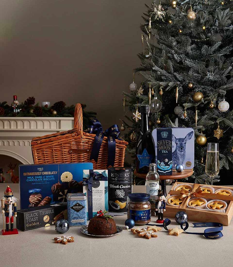 Midnight blue-themed Christmas hamper with fine teas, chocolates, truffle snacks, and festive treats near a decorated Christmas tree.
