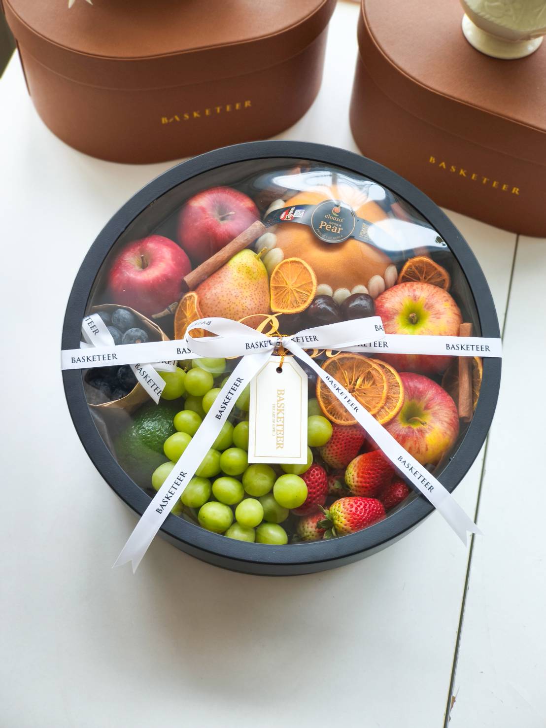 Luxury round fruit gift box with apples, grapes, strawberries, pears, and dried orange slices, wrapped with a white Basketeer ribbon.