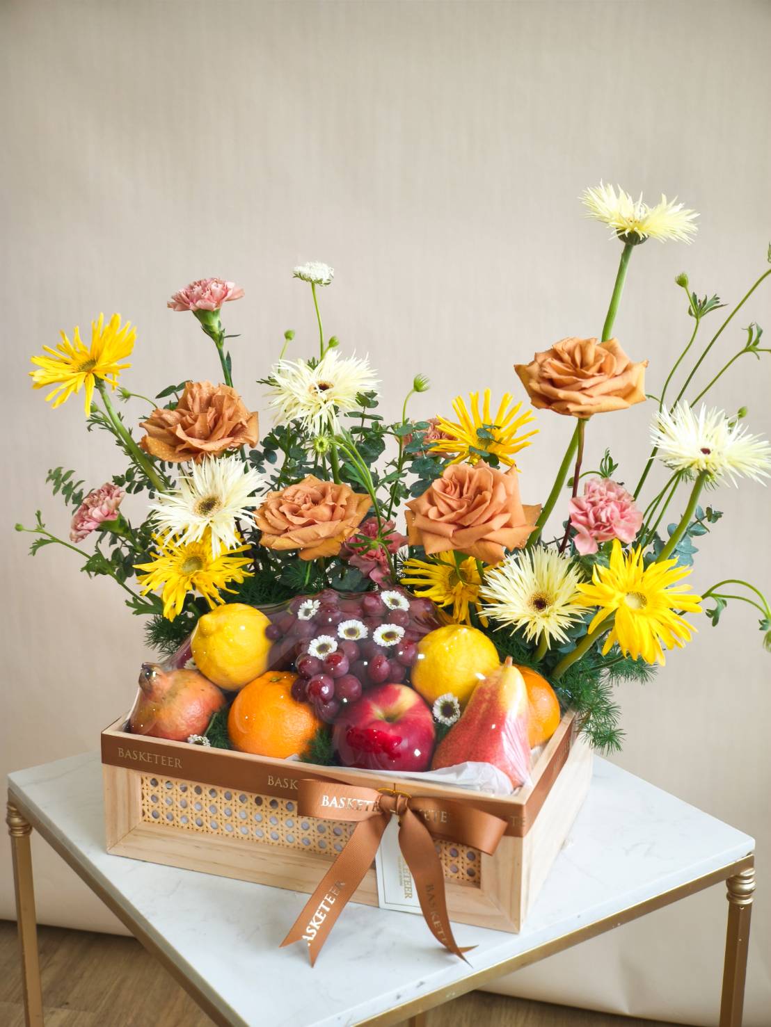 Luxury wooden basket with fresh fruits and a floral arrangement of brown roses, yellow gerberas, pink carnations, and white blooms.