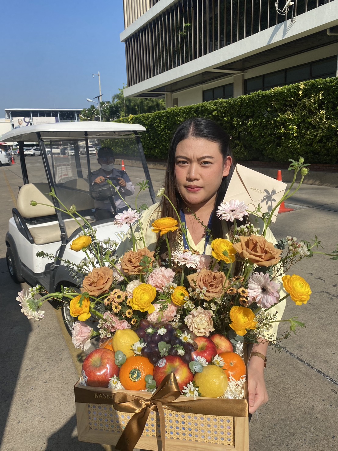 A woman holding a handcrafted fruit basket with vibrant golden and pastel flowers, fresh fruits, and a golden ribbon.