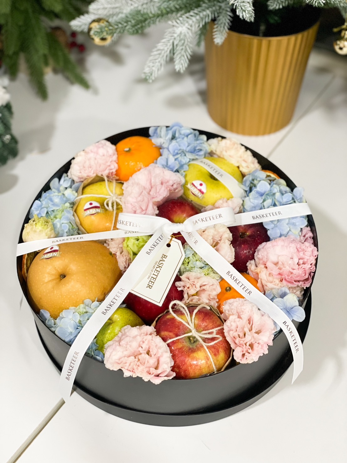 Premium round fruit box with fresh fruits, pastel hydrangeas, pink carnations, and Basketeer’s ribbon.