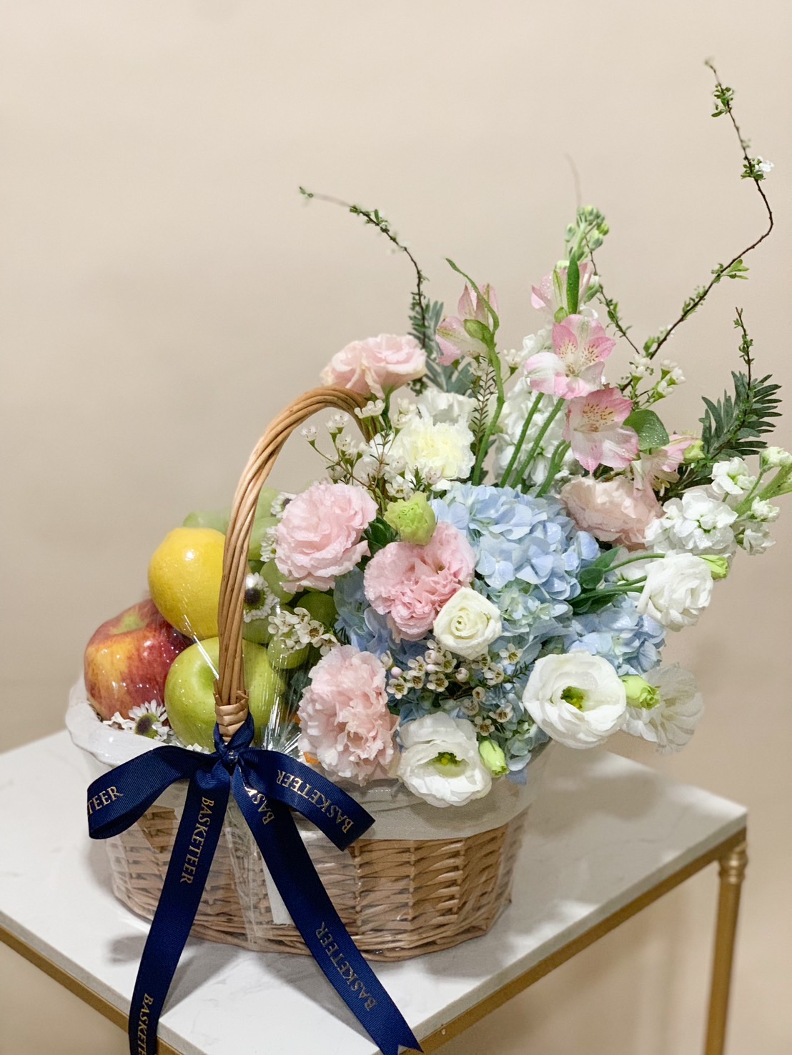 Pastel fruit basket with fresh apples, pears, hydrangeas, carnations, and lisianthus tied with a navy ribbon. Title: Elegant Pastel Floral Fruit Basket - Perfect Gift for Special Occasions Let me know if you'd like further revisions!