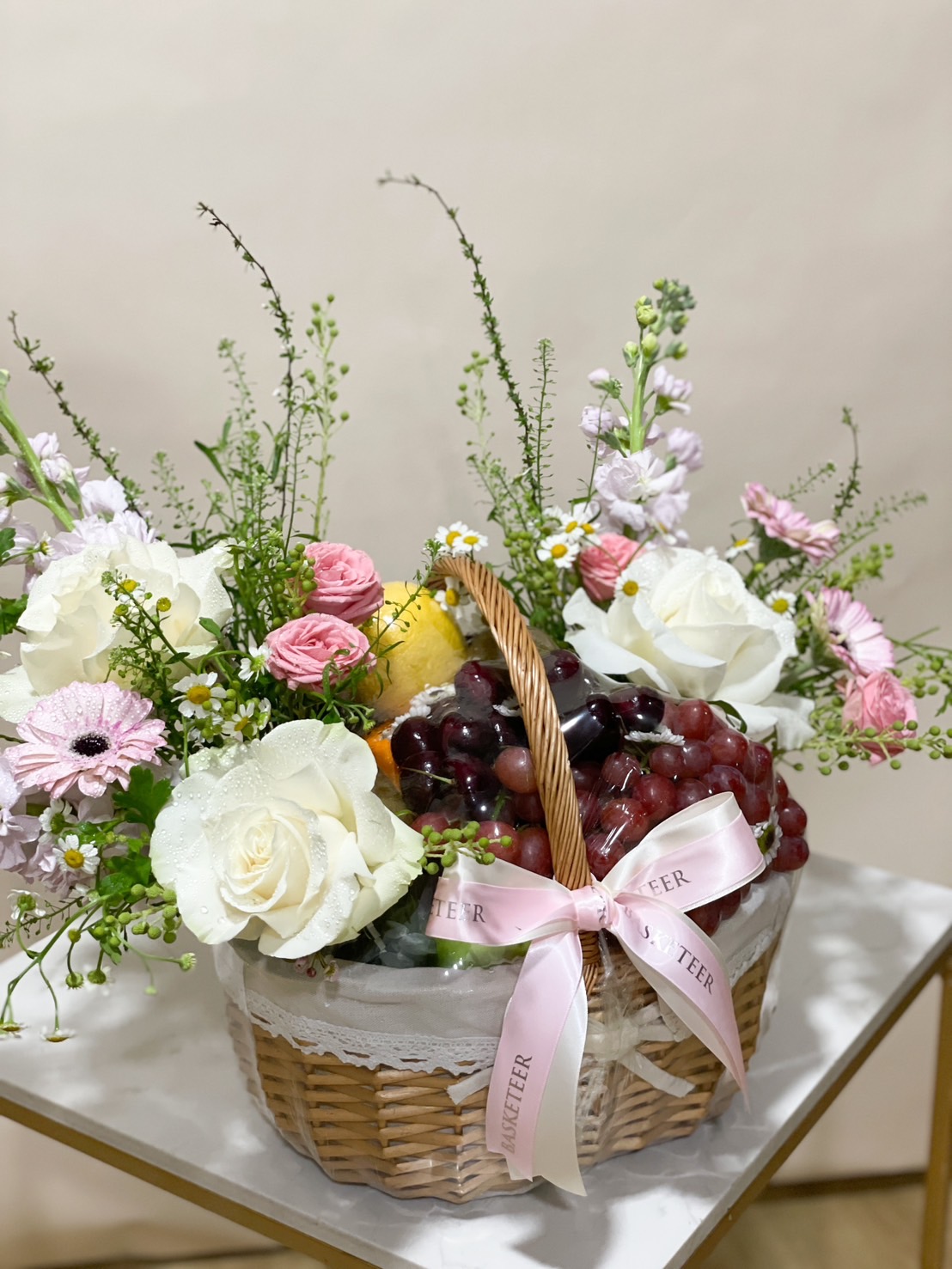 A fruit basket filled with fresh grapes, apples, roses, and daisies tied with a pink ribbon.
