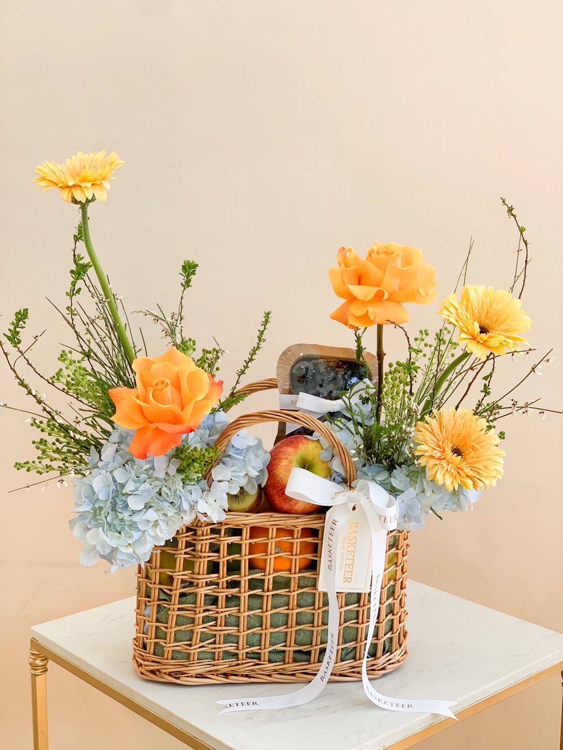 A woven basket filled with fresh apples, blueberries, and a vibrant floral arrangement of yellow gerberas, orange roses, and hydrangeas.