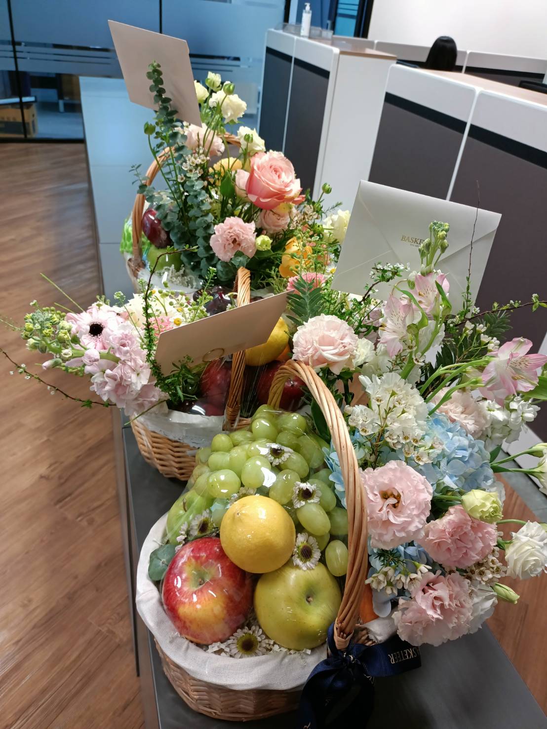 Premium fruit basket with fresh apples, grapes, lemons, and pastel flowers like roses and hydrangeas, presented elegantly for gifting.