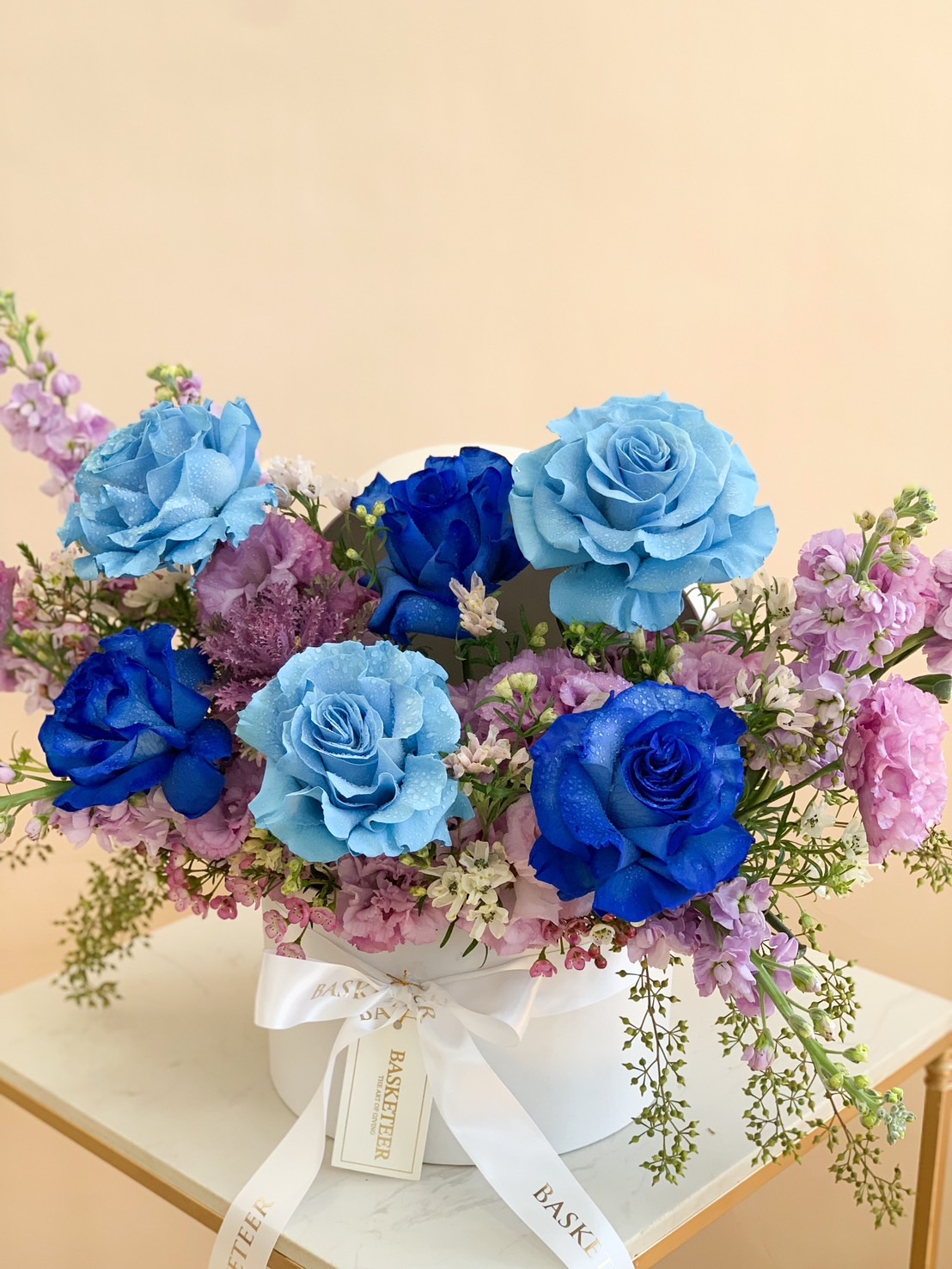 A luxurious arrangement of blue and lavender roses with purple and green accents in a white hatbox tied with a Basketeer ribbon.