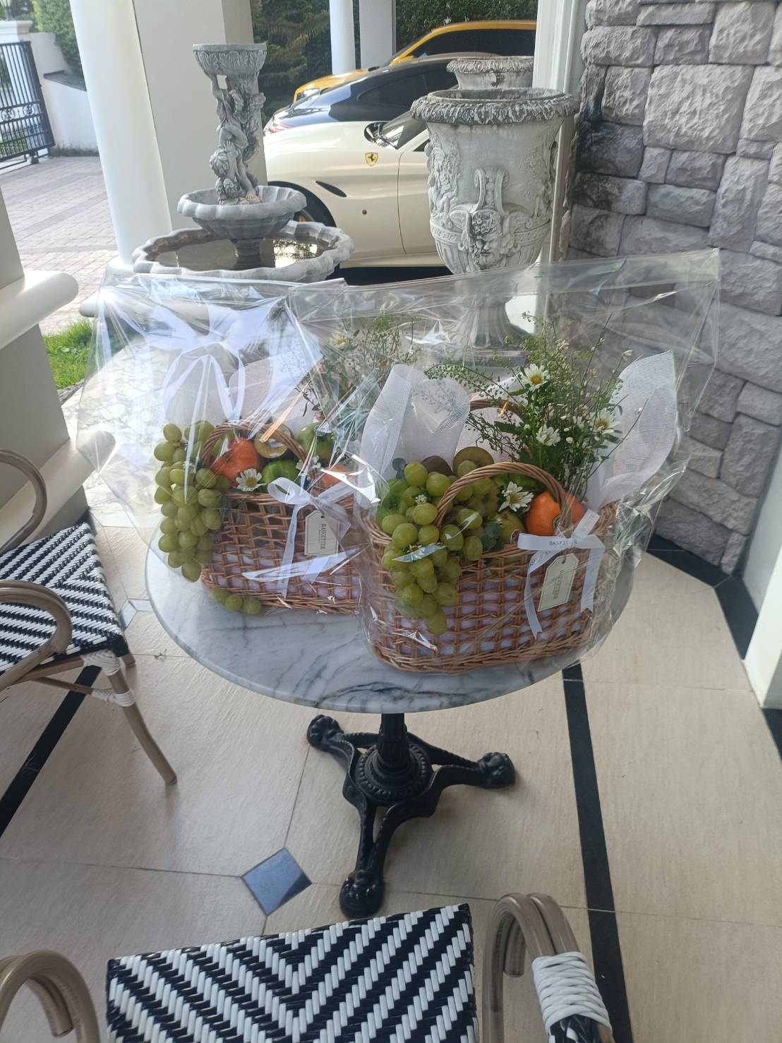 Two woven fruit baskets filled with fresh grapes, oranges, and flowers, wrapped in clear gift packaging and placed on a marble table.