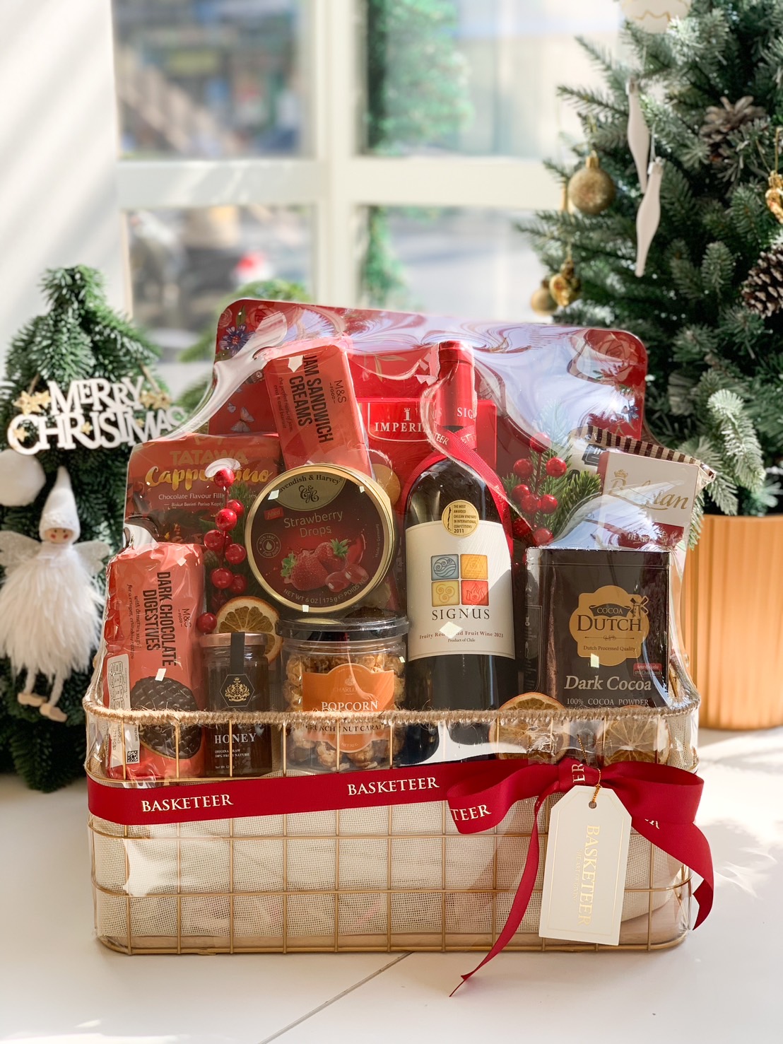 Luxurious gift hamper filled with wine, dark cocoa, honey, biscuits, and gourmet snacks, elegantly presented in a golden basket with a red Basketeer ribbon.