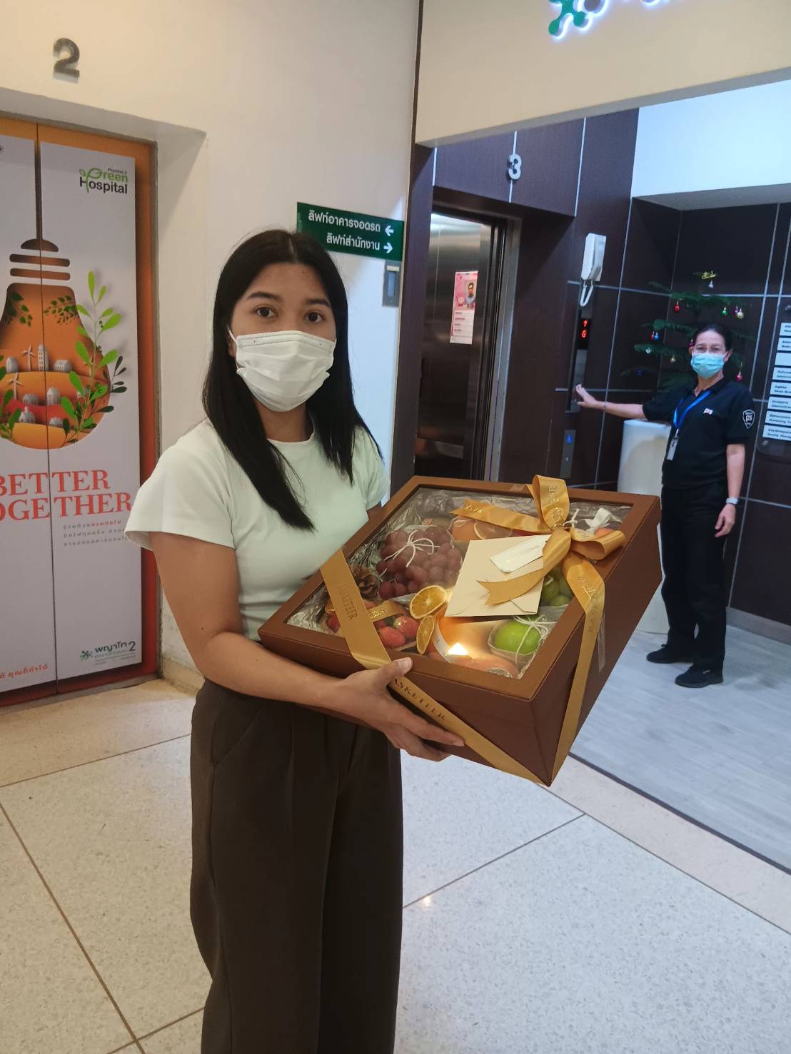 A fresh fruit gift box with apples, grapes, and oranges, adorned with golden ribbons, held by a customer at a hospital.