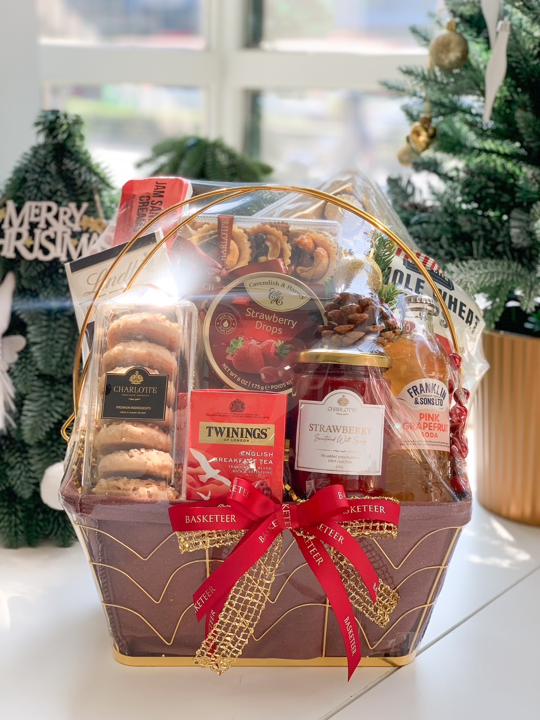 A luxurious gift basket filled with cookies, strawberry drops, tea, jam, and pink grapefruit soda, adorned with a red Basketeer ribbon and festive decorations.