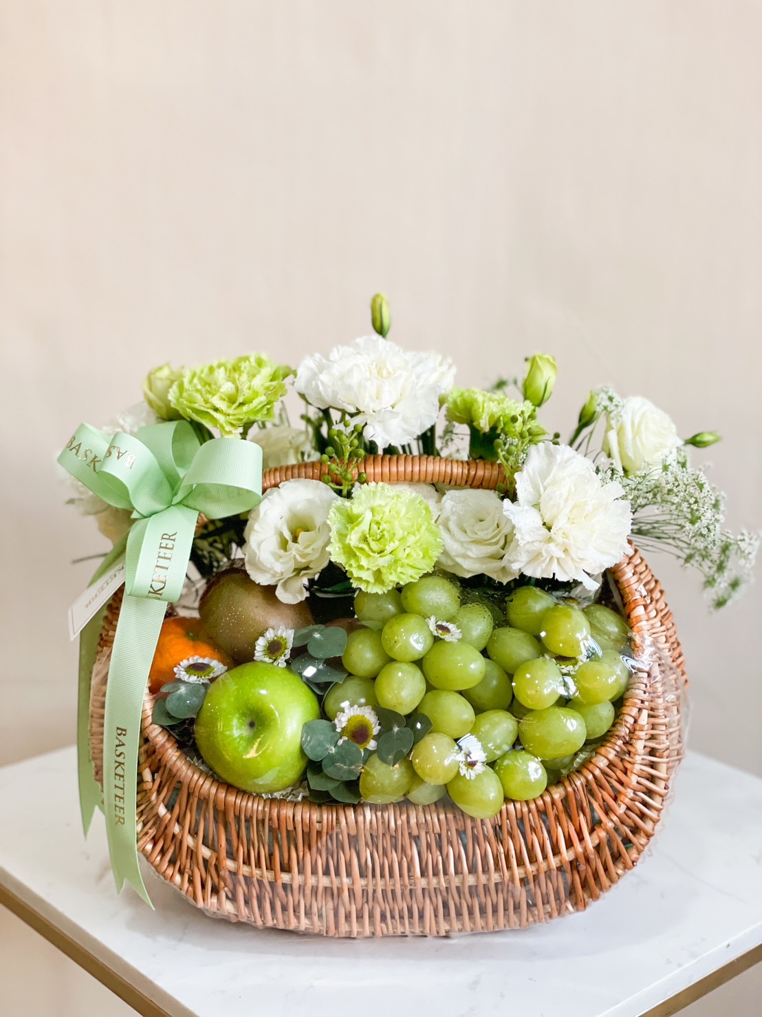 The Fresh Harmony Fruit Basket is a wicker basket filled with green grapes, a green apple, kiwi, and an orange, adorned with delicate white and green flowers. A green ribbon with text graces the basket, which rests on a white surface against a light background.
