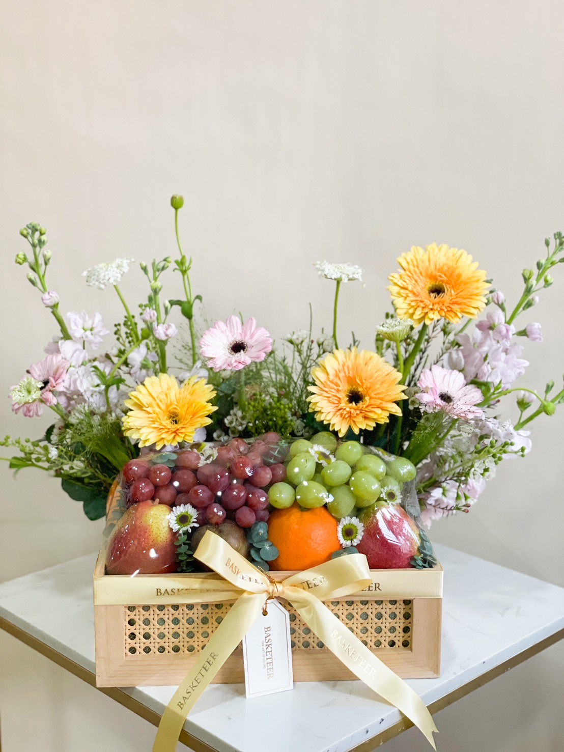 Wooden rattan basket with fresh oranges, grapes, apples, and a pastel pink and yellow flower arrangement.