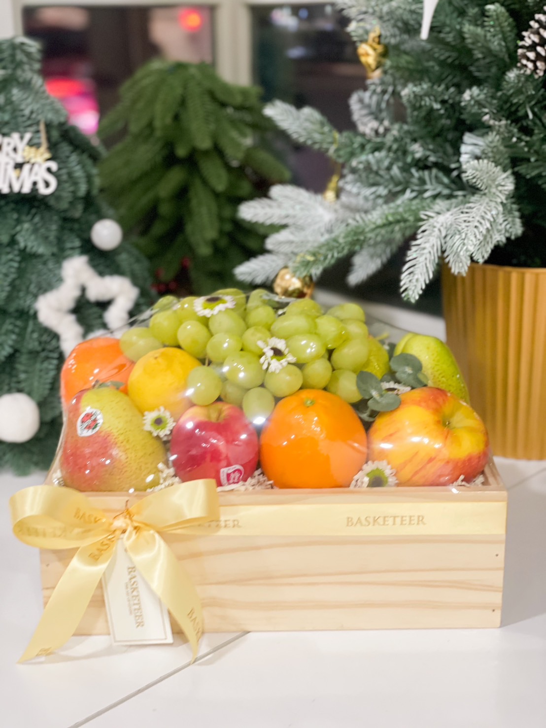 A wooden crate filled with fresh fruits, including green grapes, apples, oranges, and pears, adorned with a golden ribbon and floral accents.
