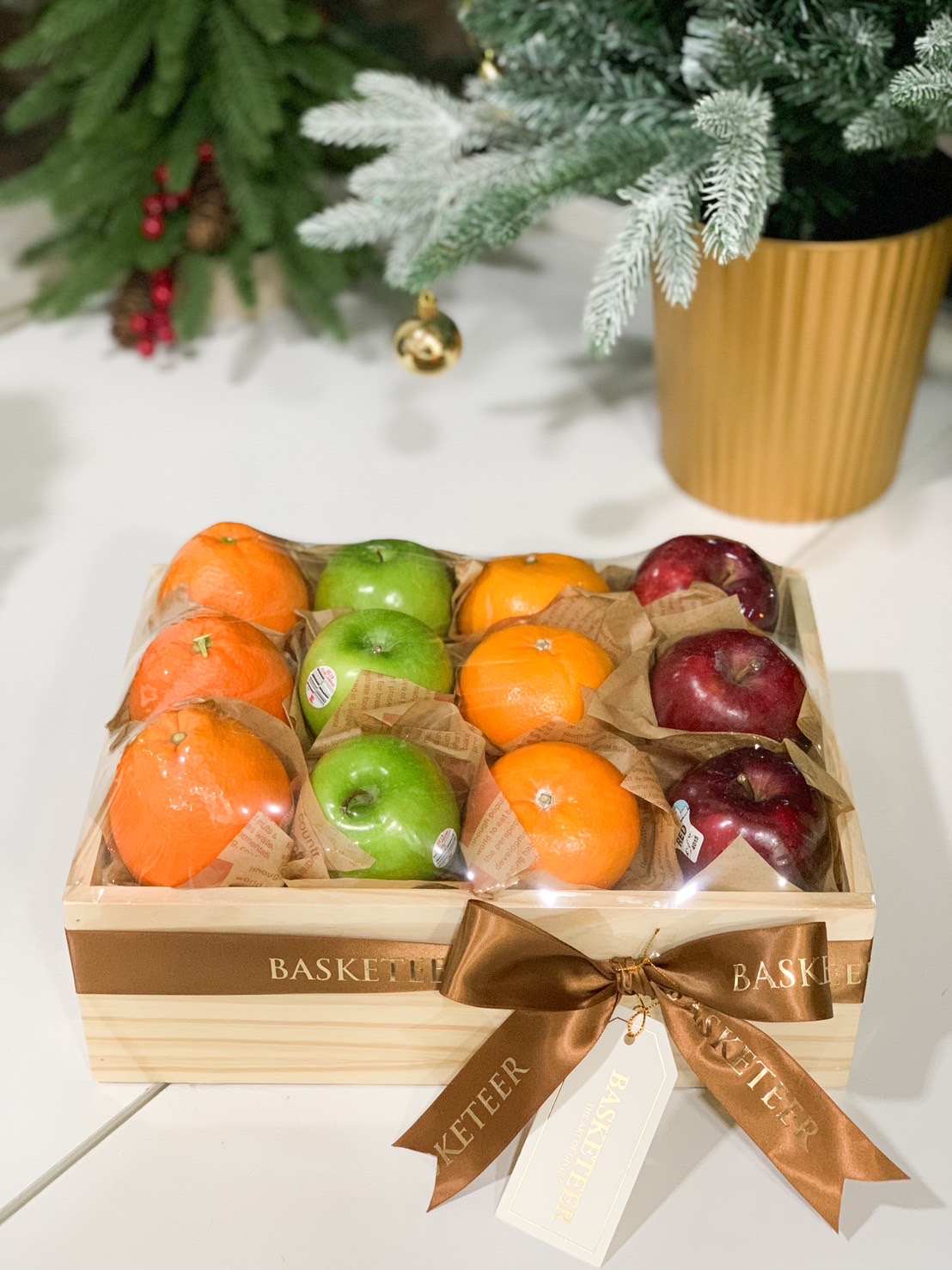 Fresh fruit gift box featuring red apples, green apples, and oranges arranged in a rustic wooden box with a brown ribbon.