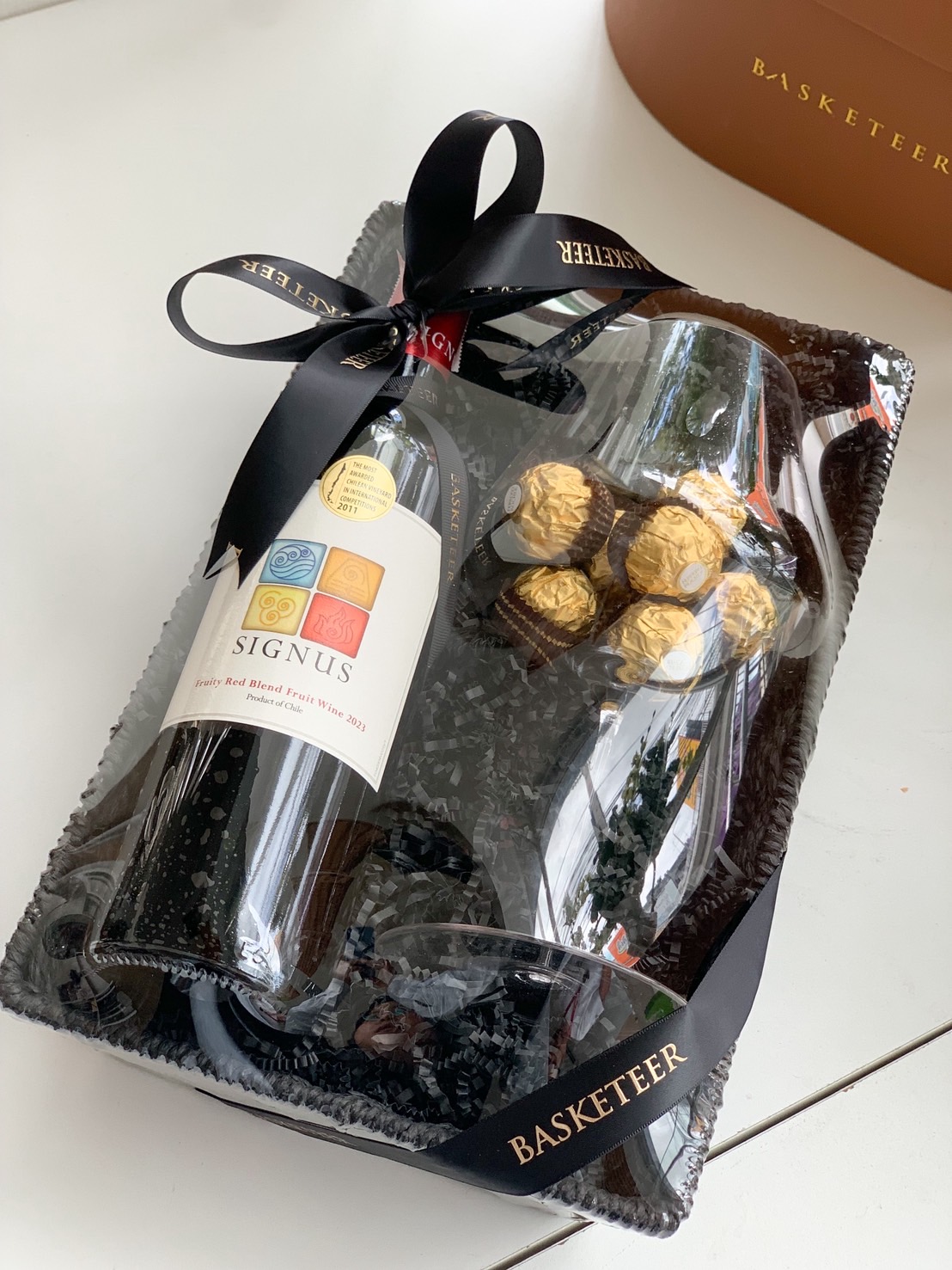 Premium wine and chocolate gift basket with Signus red wine, Ferrero Rocher, and wine glasses in black basket.