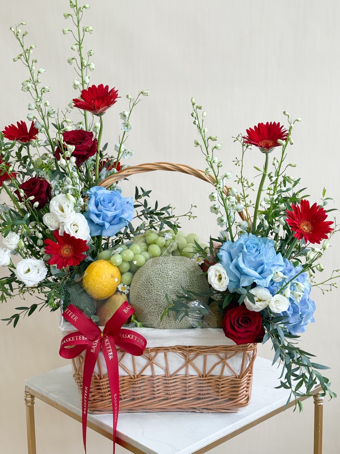 Luxury fruit basket with fresh melon, grapes, and colourful flowers including red roses, blue roses, and gerberas.