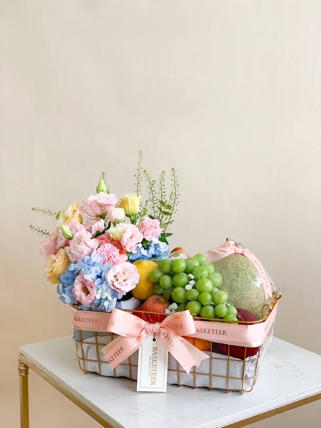 Premium fruit basket with Japanese melon, grapes, apples, and pastel flowers including pink lisianthus and blue hydrangea.