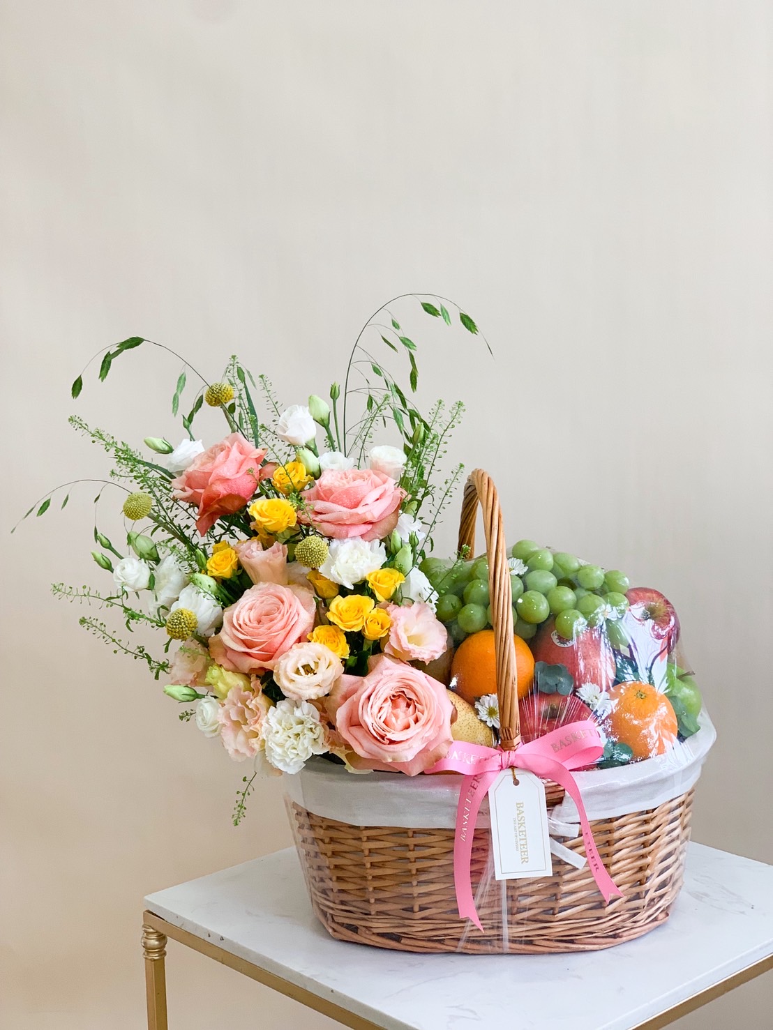 Deluxe fresh fruit basket with pink roses, white lisianthus, and yellow spray roses in premium woven basket.