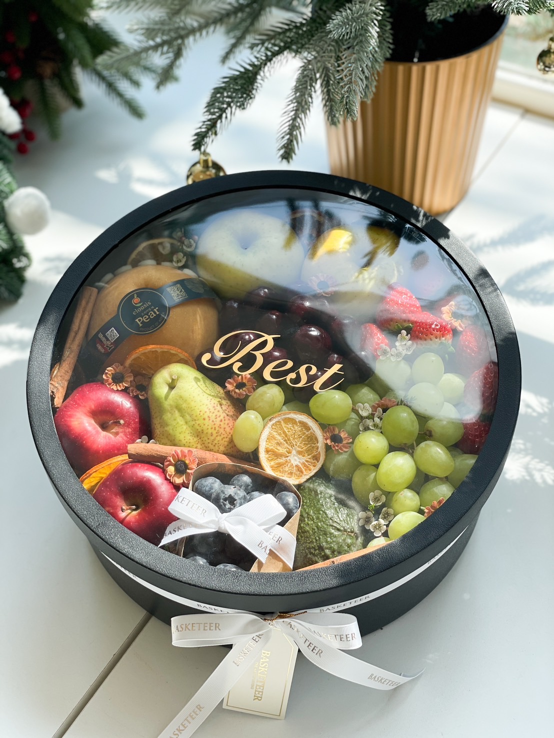 Luxury fresh fruit box with apples, pears, grapes, strawberries, and dried oranges in an elegant black round box, adorned with white ribbon and 'Best' text on the lid.