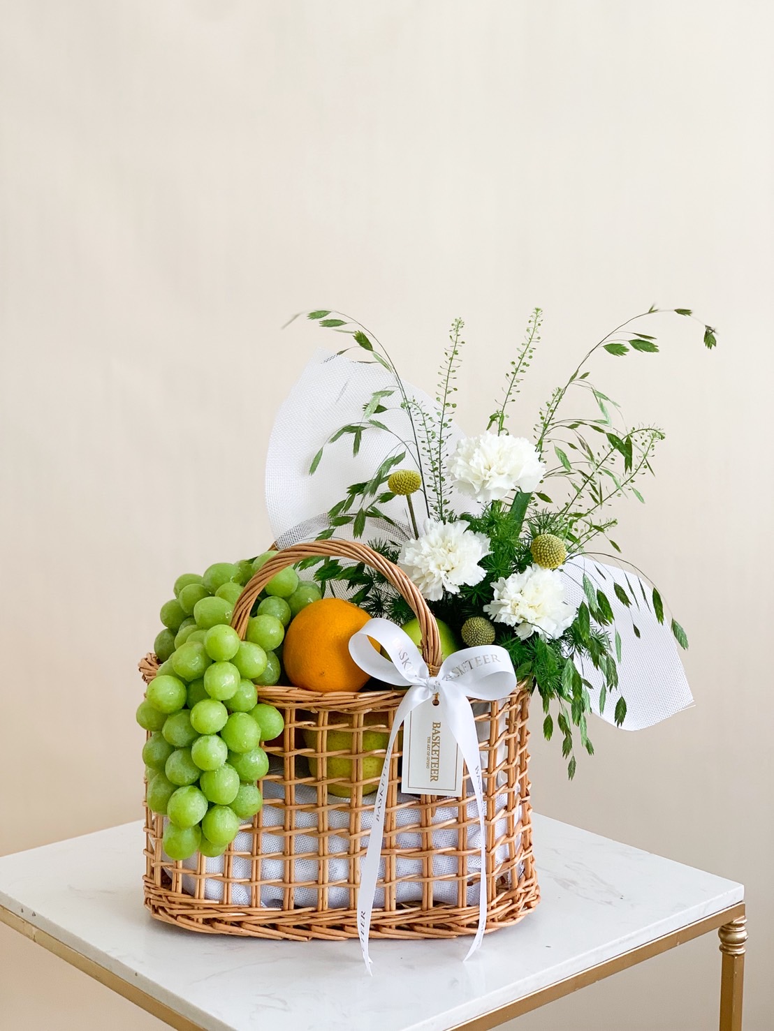 Elegant wicker fruit basket with green grapes, orange, and white flower arrangement with a white ribbon.