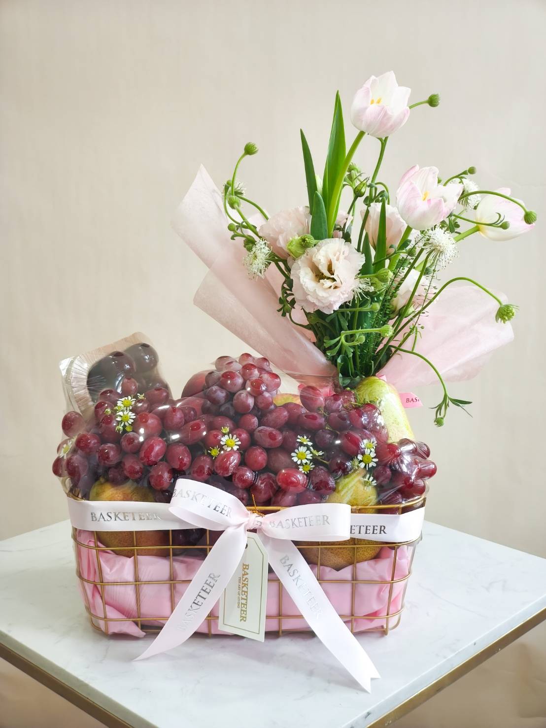 Luxury fruit basket with fresh red grapes, assorted fruits, and pink tulips with lisianthus flowers, wrapped with ribbon.