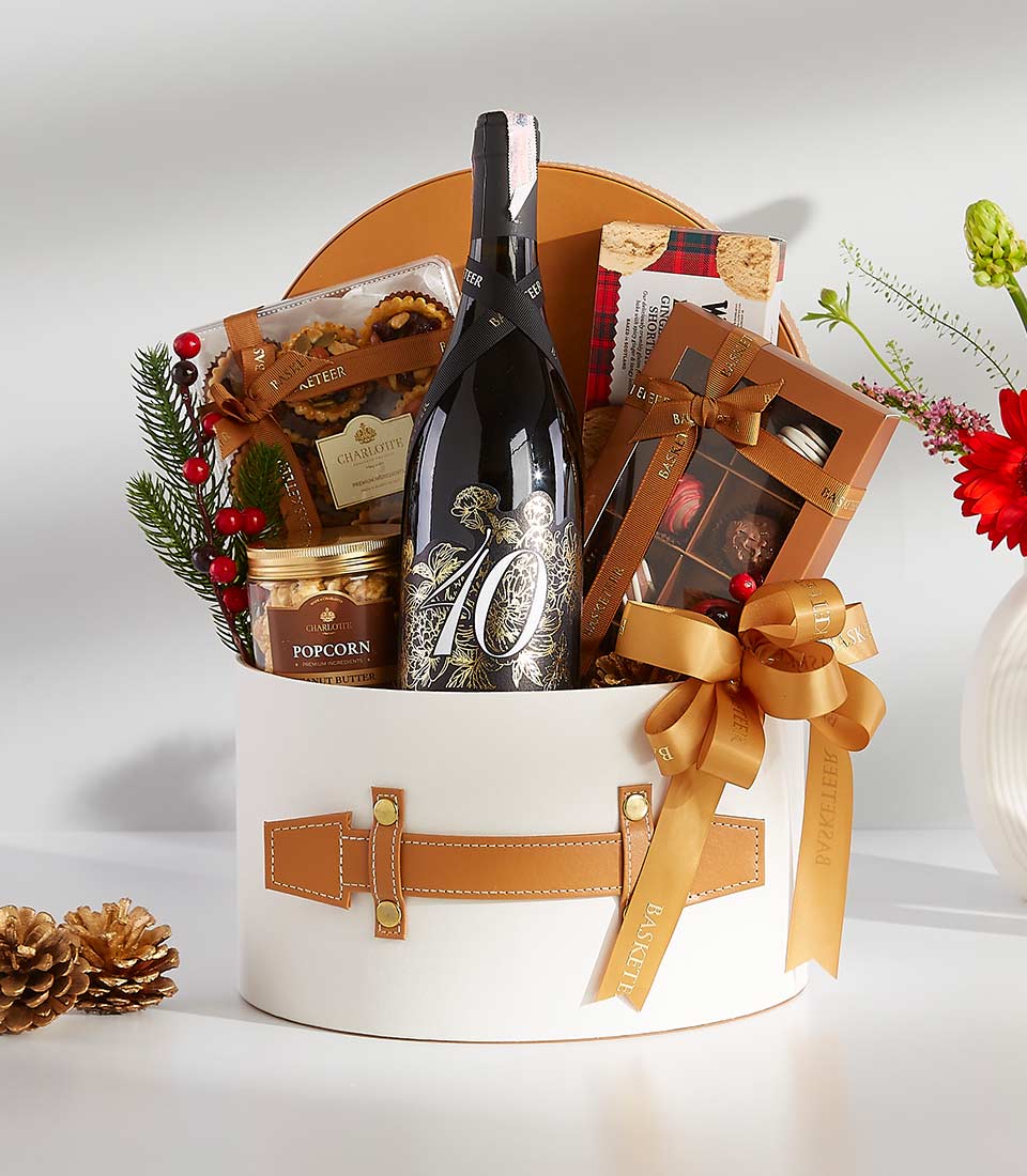 A white gift box with a brown leather handle filled with various gourmet treats, including a bottle of wine, chocolates, cookies, popcorn, and nuts.