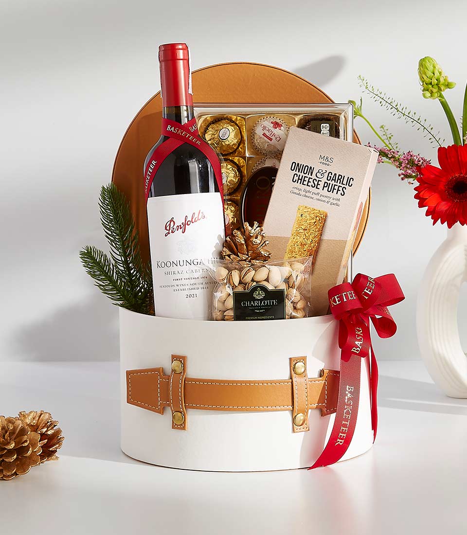 A white gift box with a brown leather handle filled with various gourmet treats, including a bottle of wine, chocolates, cookies, popcorn
