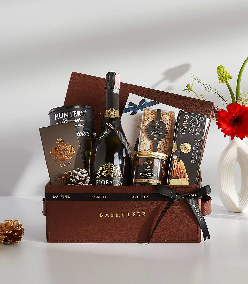 A brown leather gift box filled with various gourmet treats, including a bottle of champagne, caviar, chocolates, popcorn, and other snacks.