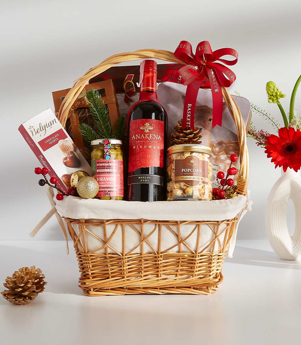 A wicker basket overflowing with festive treats, including a bottle of red wine, gourmet chocolates, popcorn, and other delicious snacks.