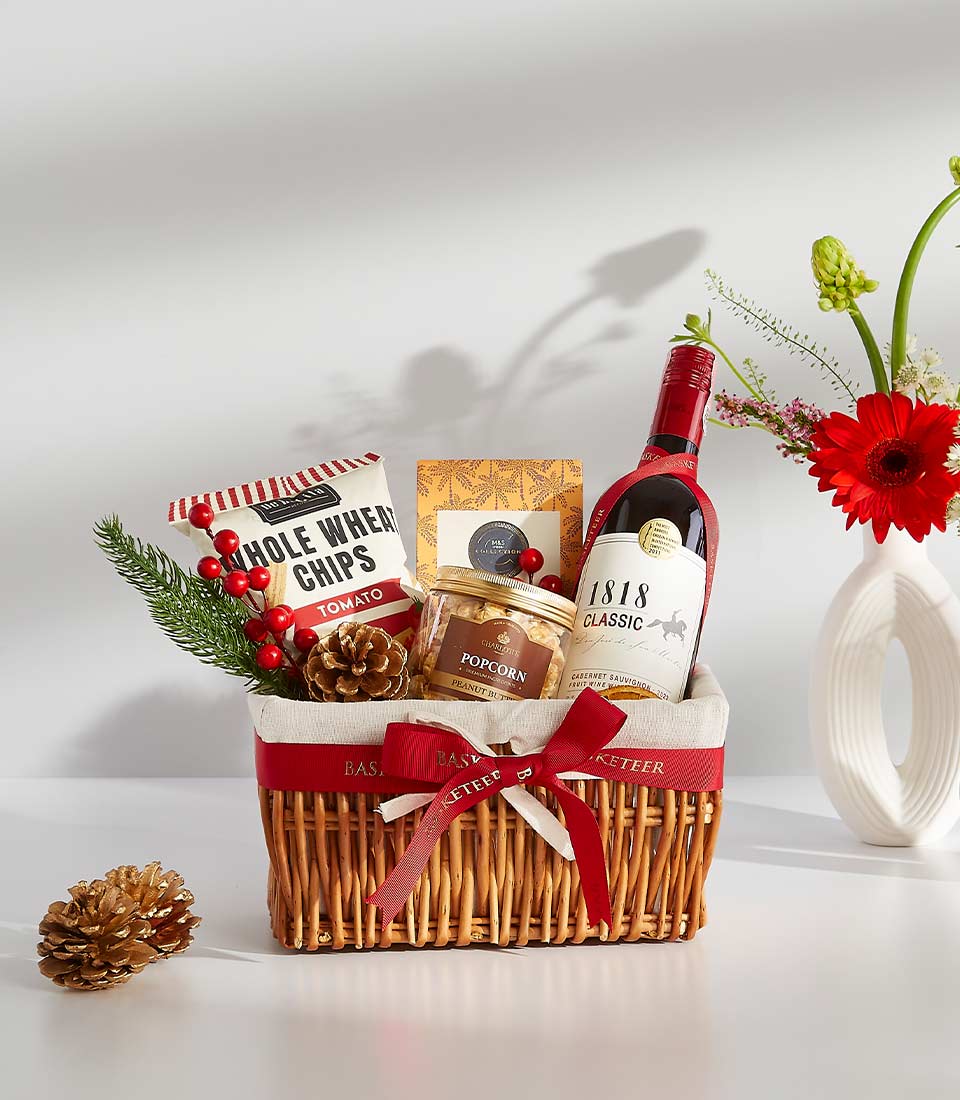 A wicker basket overflowing with festive treats, including a bottle of red wine, gourmet chocolates, popcorn, and other delicious snacks.