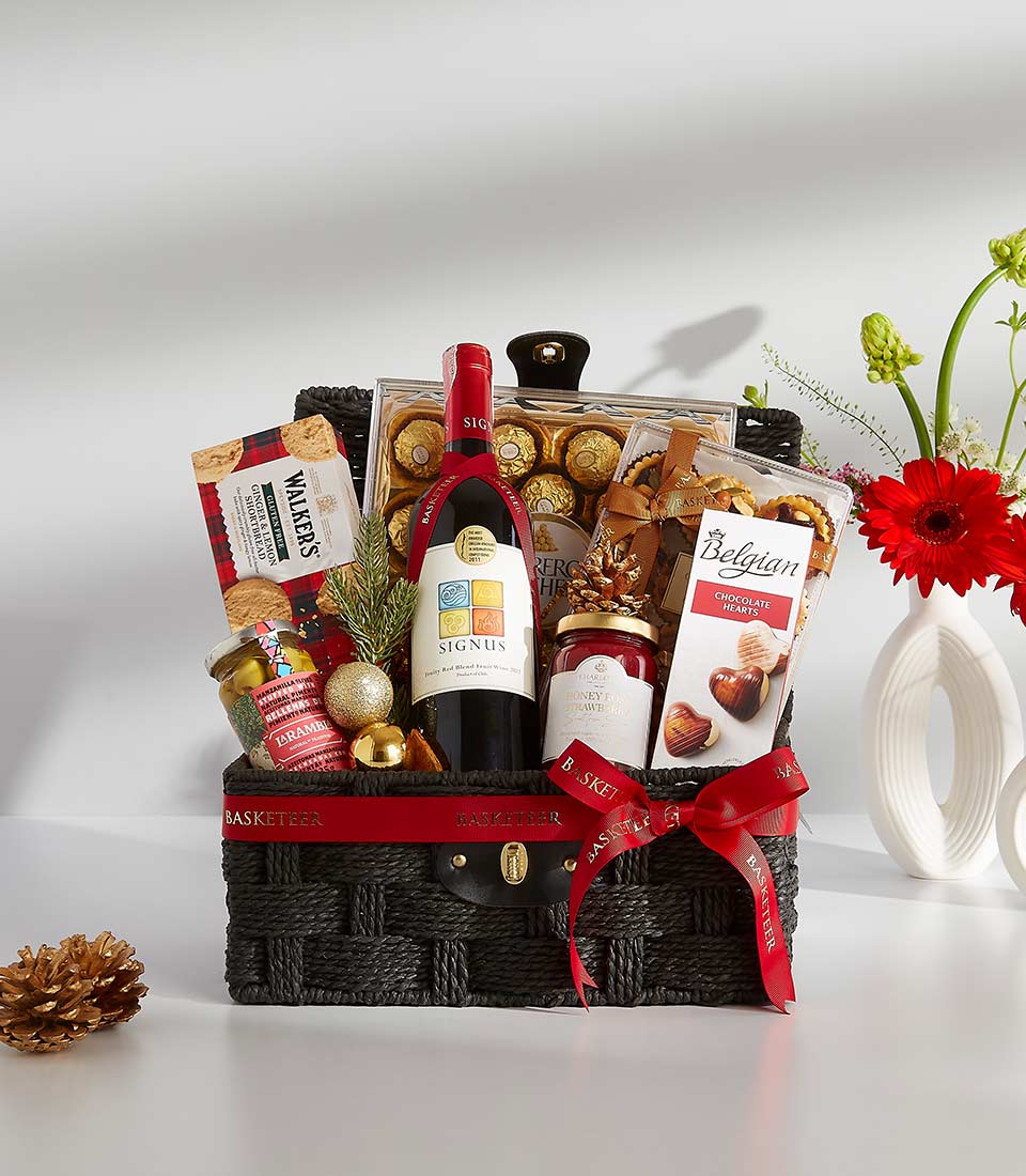 A black wicker basket overflowing with festive treats, including a bottle of rich red wine, a box of assorted chocolates, and other goodies.