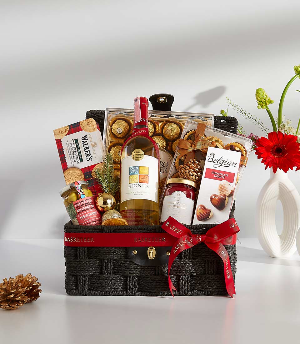 A black wicker basket overflowing with festive treats, including a bottle of rich red wine, a box of assorted chocolates, and other goodies.