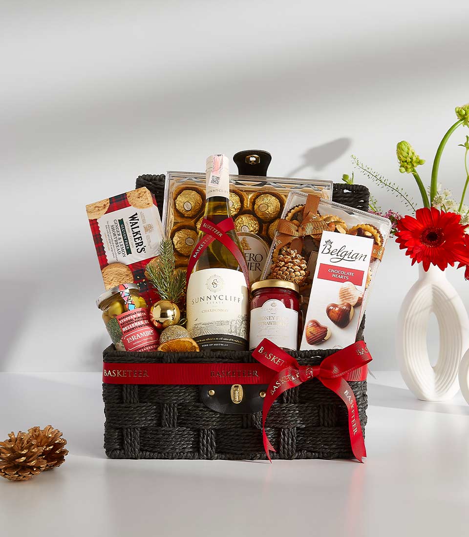 A black wicker basket overflowing with festive treats, including a bottle of rich red wine, a box of assorted chocolates, and other goodies.