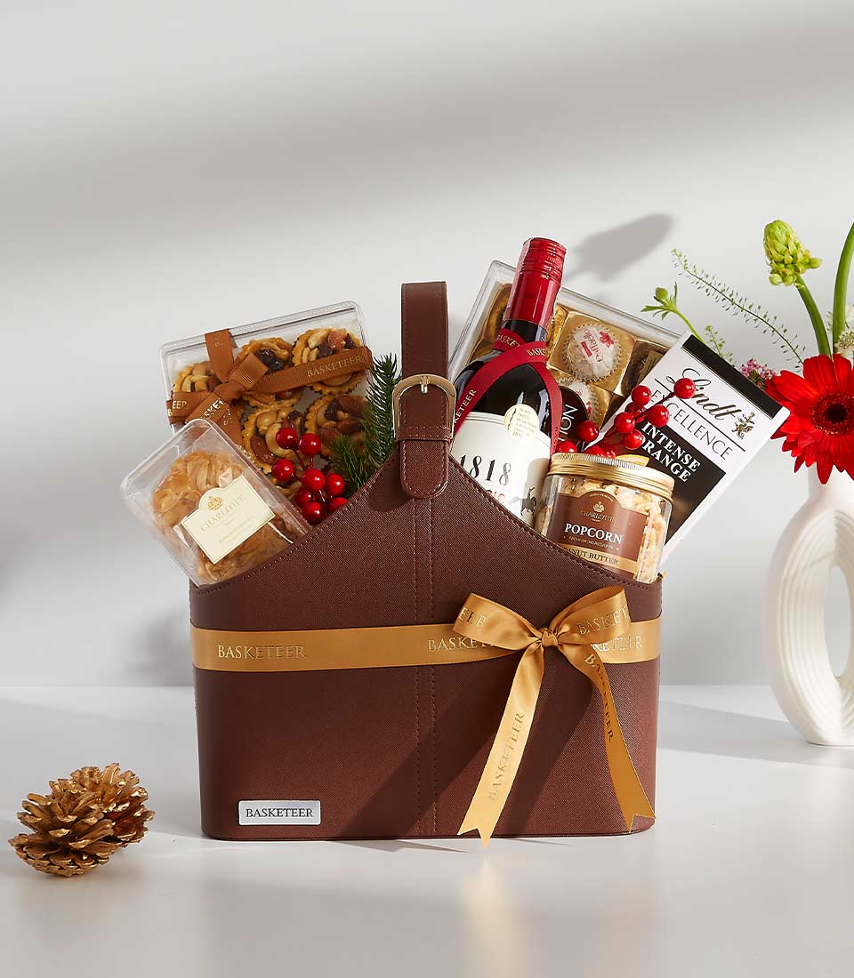 A brown leather basket filled with a variety of gourmet treats, including a bottle of wine, chocolates, cookies, and popcorn.