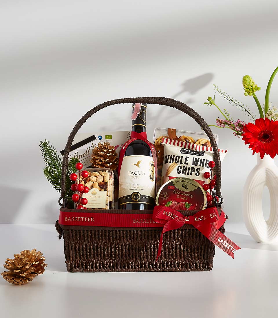 A brown wicker basket filled with a variety of gourmet treats, including a bottle of wine, chocolates
