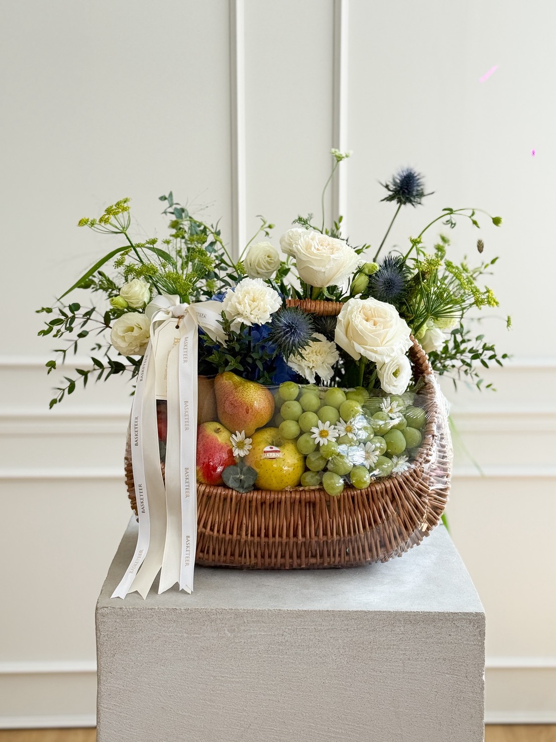 A refined wicker hamper brimming with fresh fruit, lush white roses, blue thistles, and elegant greenery, adorned with a white ribbon.