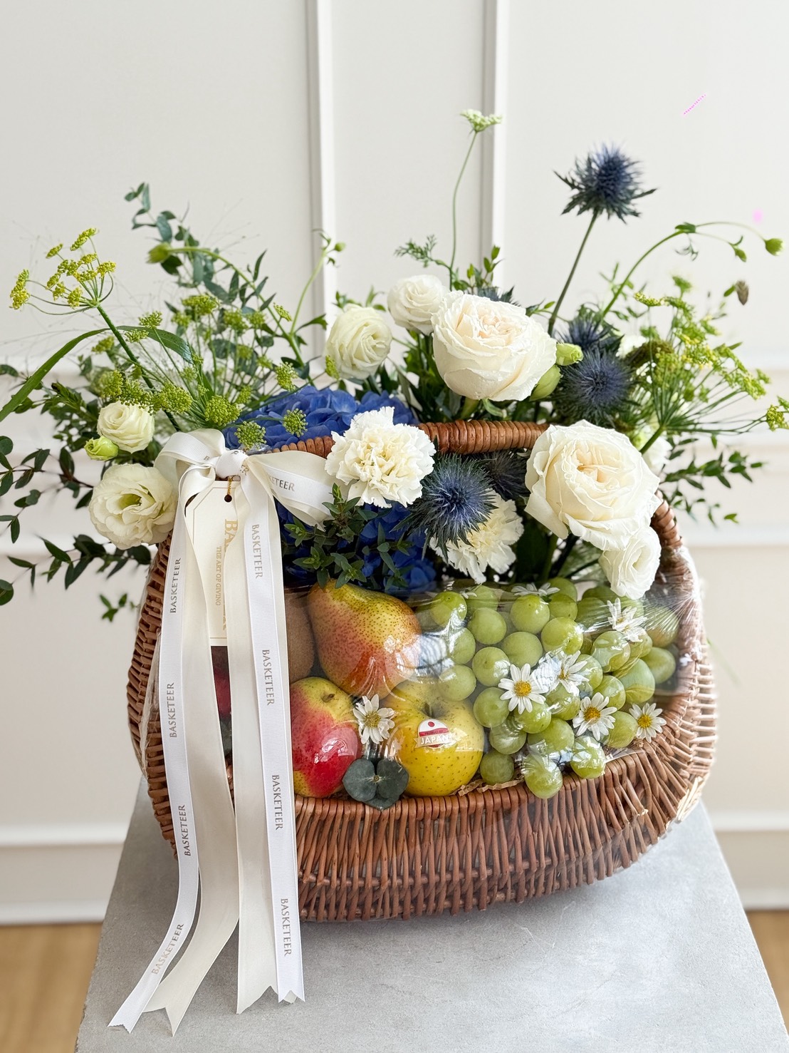 An elegant wicker hamper overflows with green grapes, pears, and apples, accented by white roses, blue blooms, and lush greenery.