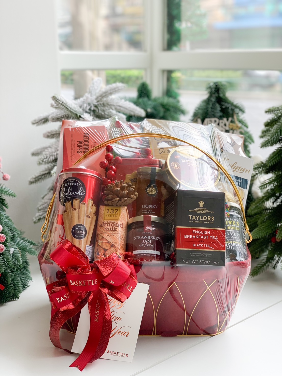 Festive gift hamper with assorted teas, jams, cookies, snacks, and a red and gold decorative ribbon.