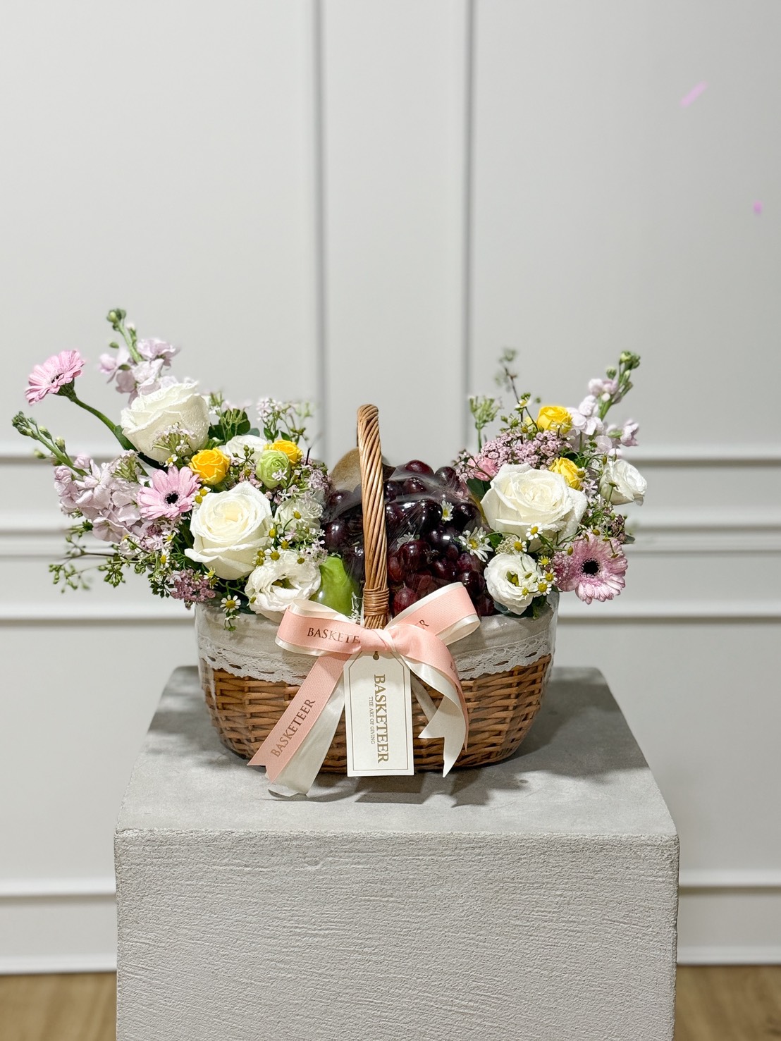 A woven gift basket filled with red grapes in the center, surrounded by white roses, yellow spray roses, pink daisies, and soft pastel flowers, decorated with pink and cream ribbons and a tag labeled “Basketeer.” The basket is placed on a concrete pedestal against a light-colored wall.