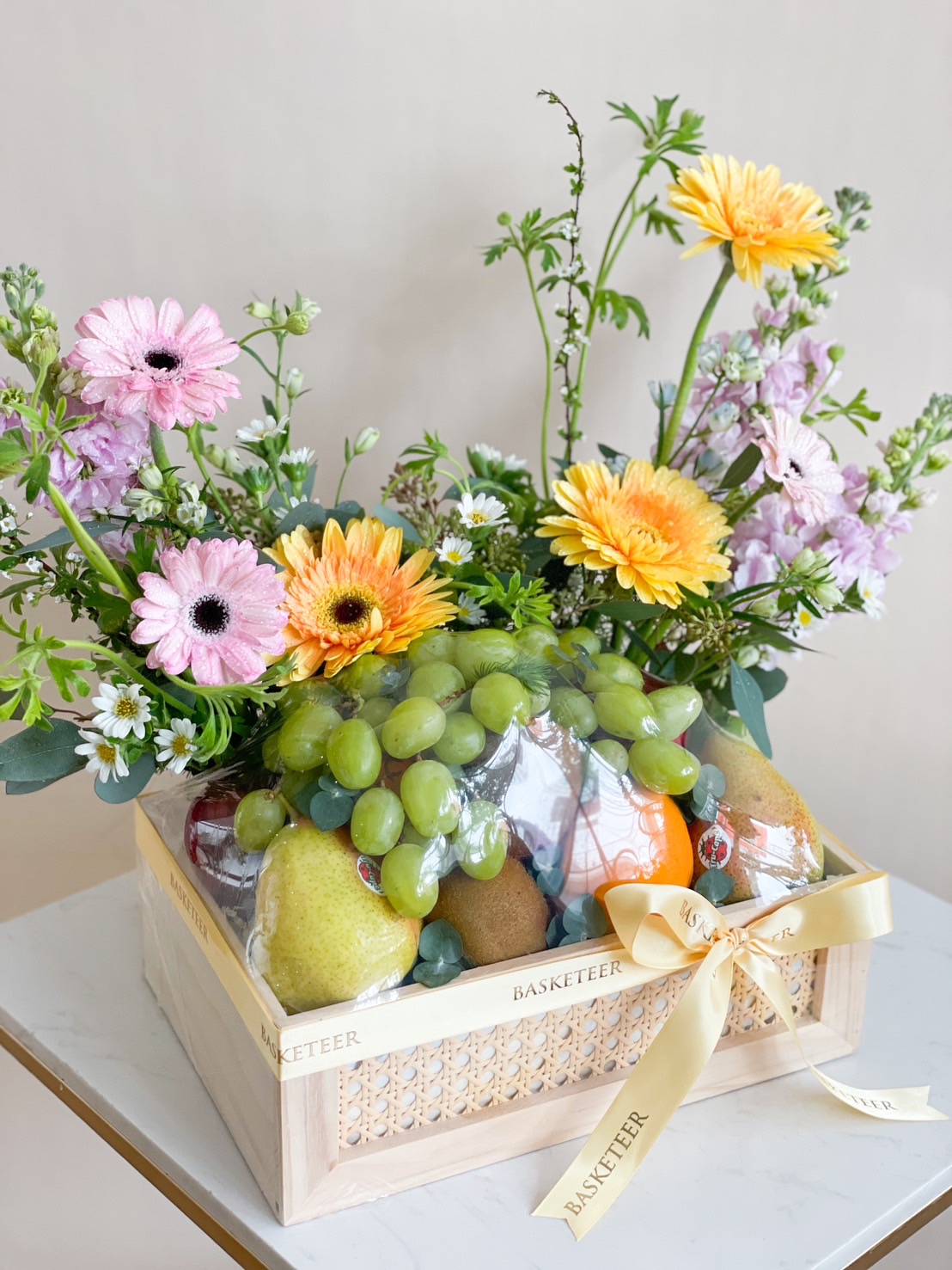 Floral fresh fruit box with grapes, kiwi, apples, citrus, and a bouquet of yellow and pink flowers.