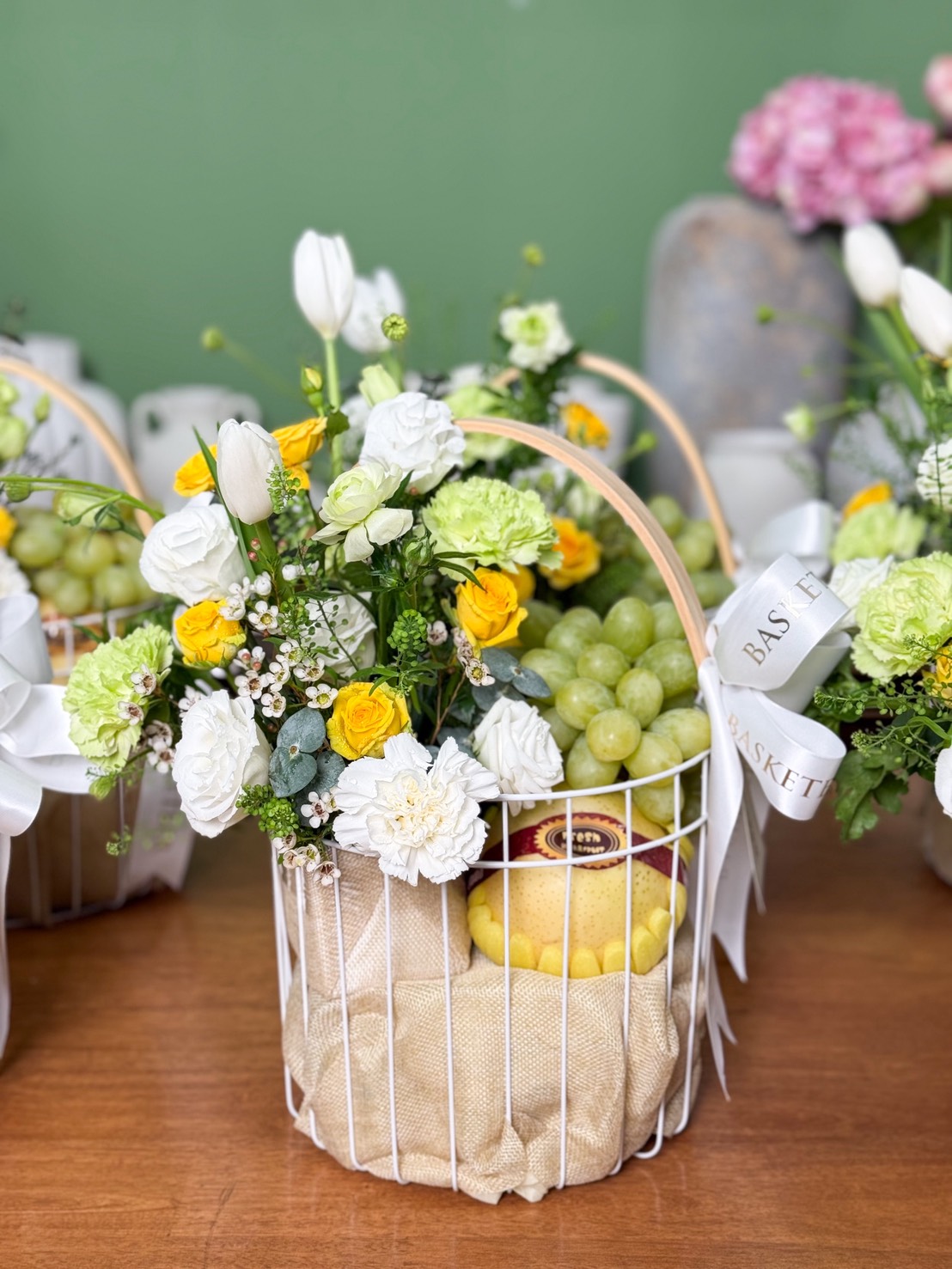 A white metal gift basket filled with green grapes and a large wrapped pear, decorated with white roses, yellow spray roses, green carnations, tulips, and small white blooms. The basket is lined with beige fabric and tied with white ribbons, placed on a wooden table with a green background.