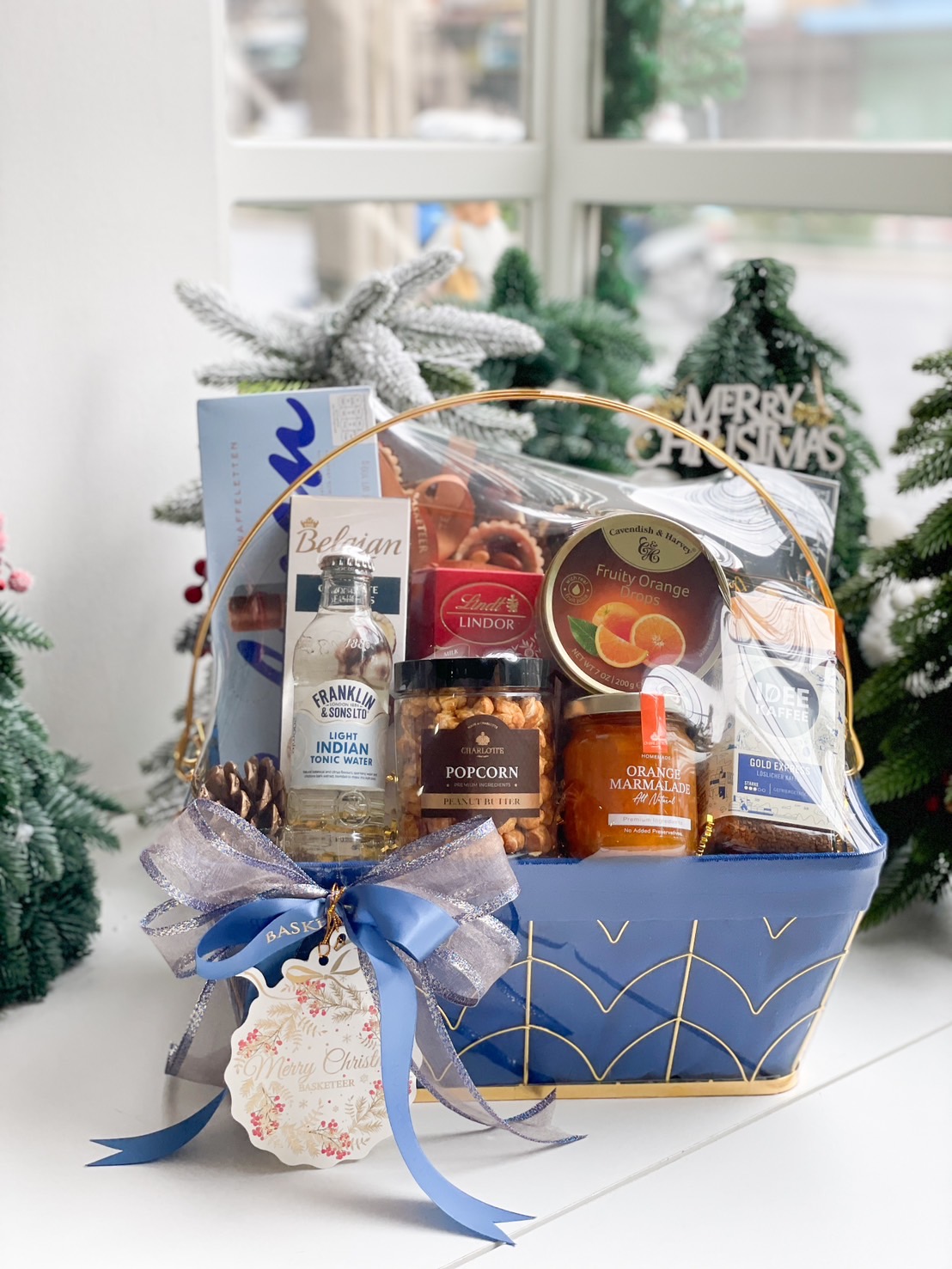 A luxurious blue gourmet gift basket with premium treats, including coffee, juice, and snacks, decorated with festive ribbons and pinecones.
