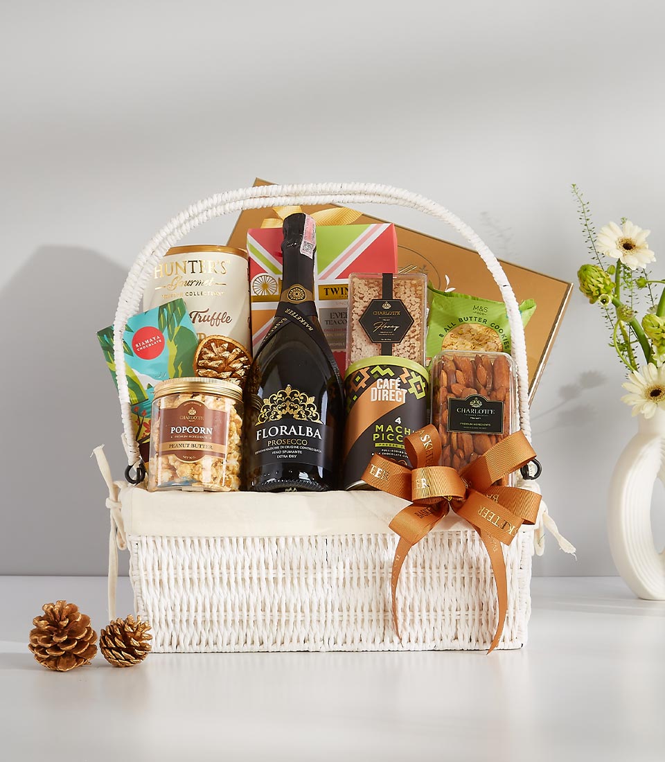 A white wicker gift basket filled with gourmet goodies including a bottle of prosecco, assorted chocolates, popcorn, nuts, and crackers.