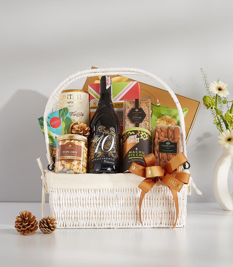 A white wicker gift basket filled with gourmet goodies including a bottle of prosecco, assorted chocolates, popcorn, nuts, and crackers.