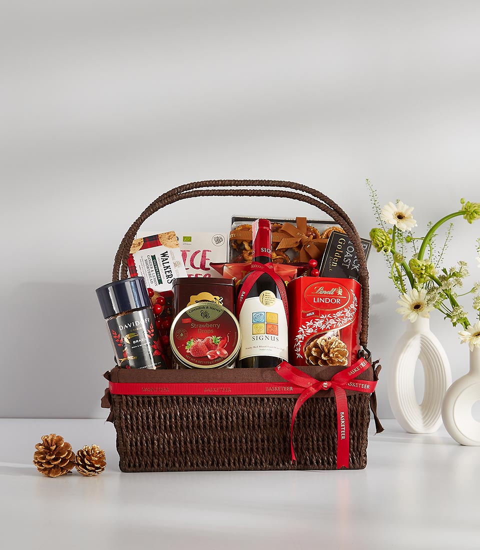 A brown wicker gift basket filled with gourmet goodies including a bottle of red wine, chocolate truffles, cookies, and other snacks.