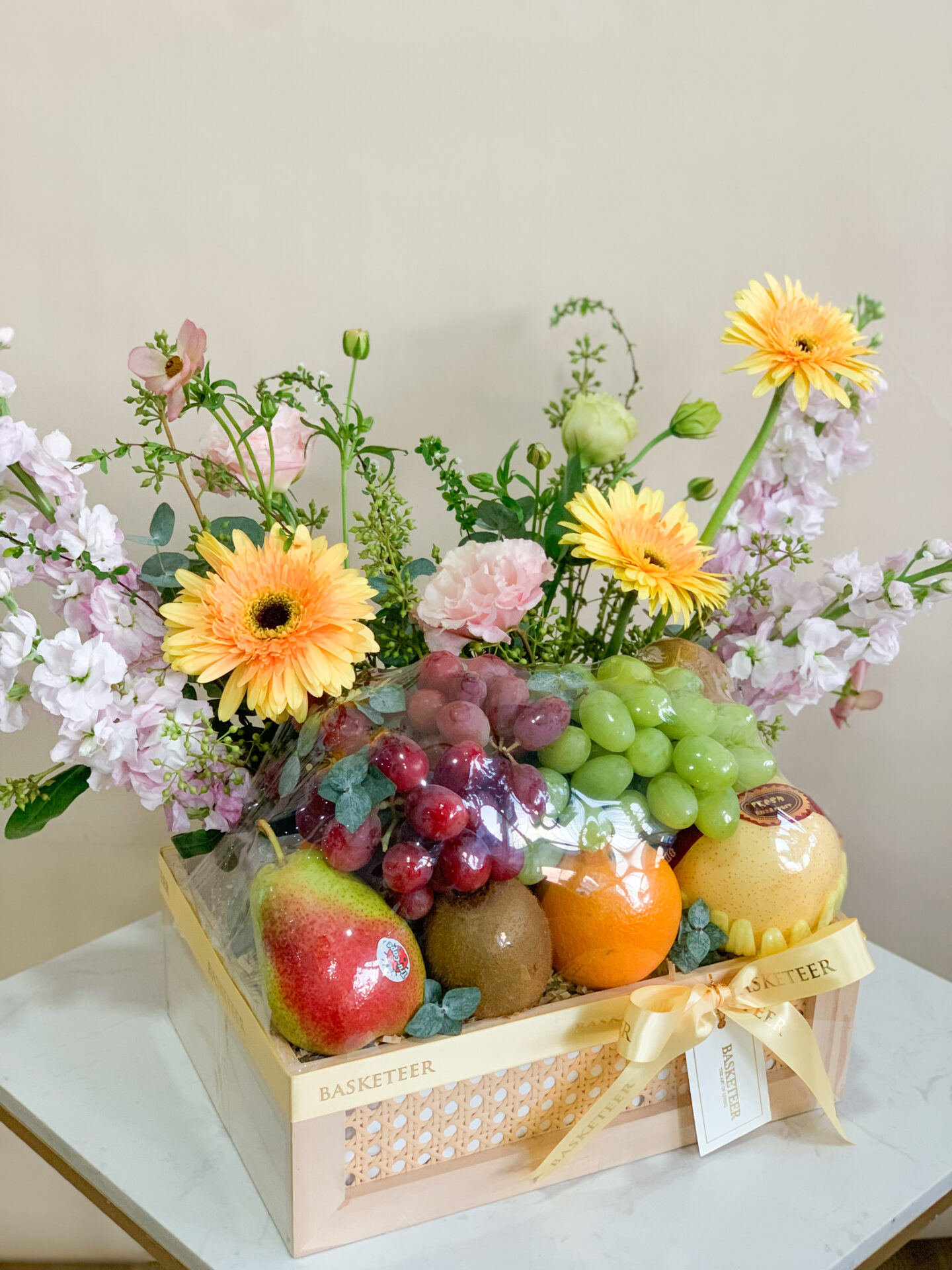 Floral fresh fruit box with grapes, kiwi, apples, citrus, and a bouquet of yellow and pink flowers.