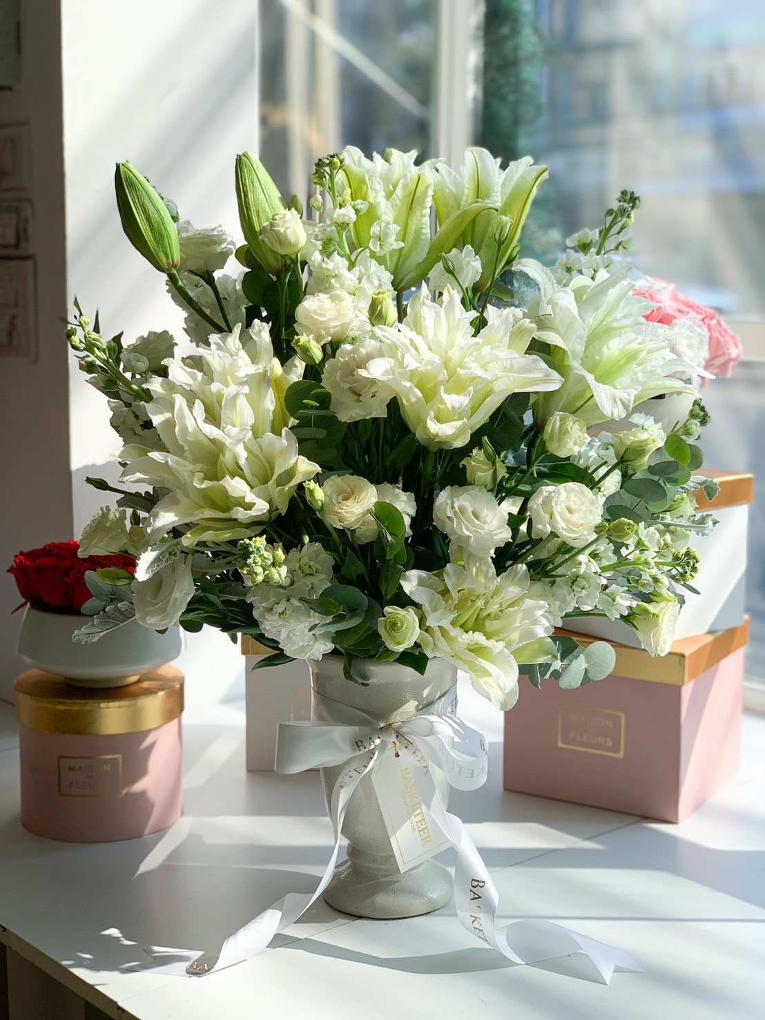 A luxurious white floral arrangement featuring elegant lilies, roses, and greenery, beautifully arranged in a ceramic vase with a Basketeer ribbon, set in a bright, modern space.