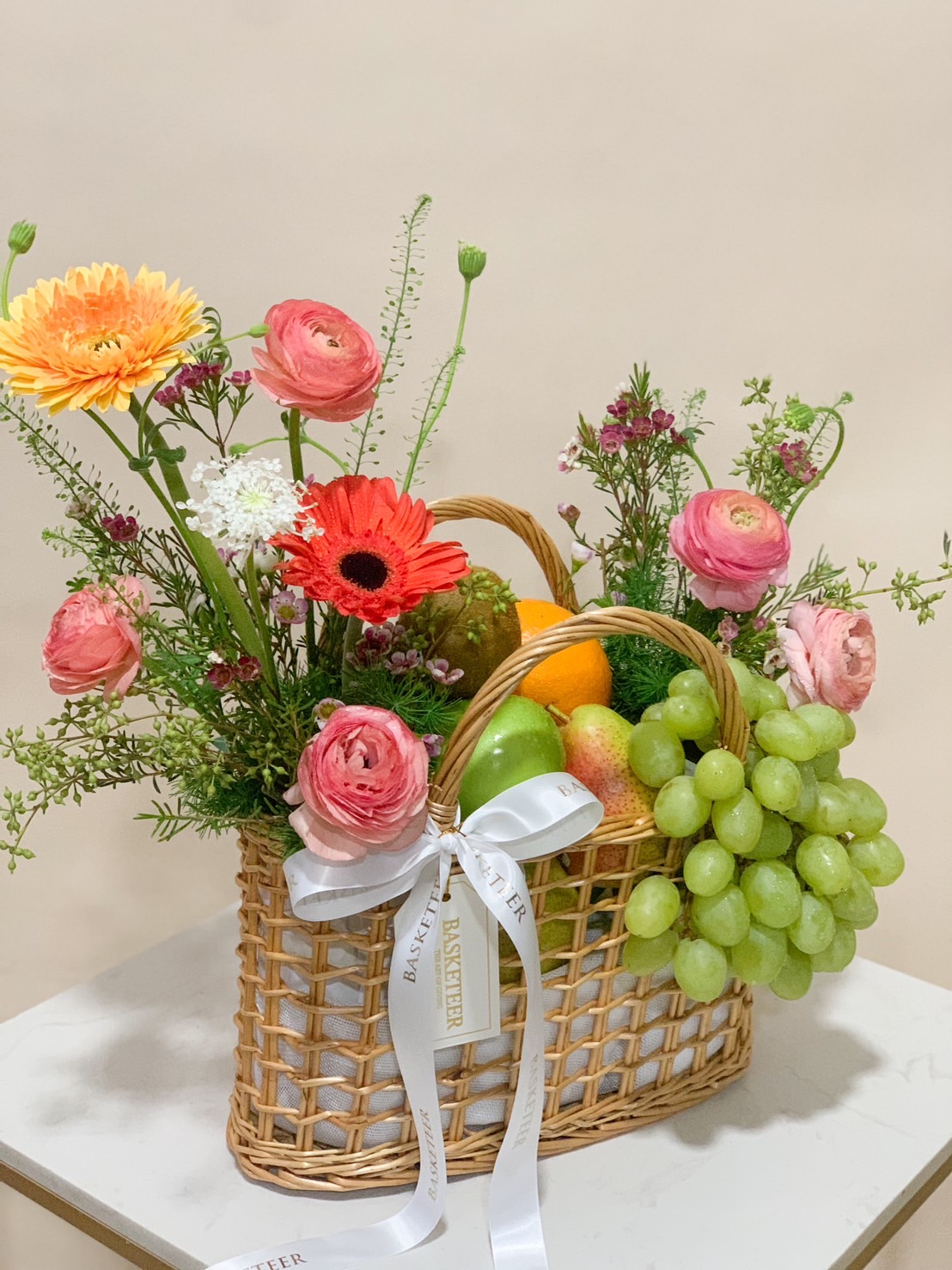 A wicker basket filled with orange and yellow flowers, green grapes, and assorted fruits like green apples, pears, and oranges. Above the basket, more fruits are floating against a neutral background. Some fruits are placed around the basket.